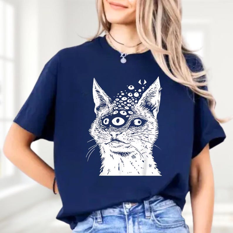 以下が含まれることがあります： 複数の目を持つ猫の白いイラストが特徴のネイビーブルーのTシャツ。グラフィックデザインは詳細で様式化されています。このカジュアルなTシャツは、ユニークなデザインで、普段使いに最適です。