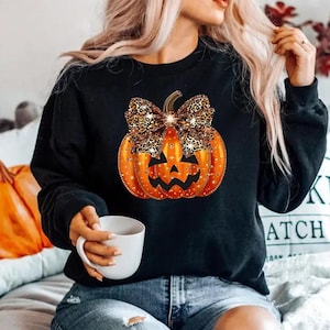 May include: A black long-sleeve sweatshirt featuring a central graphic of an orange jack-o'-lantern pumpkin adorned with a large leopard print bow. The pumpkin design includes sparkling star accents and a classic carved face with triangular eyes.