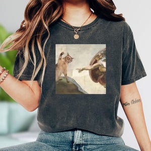 A woman wearing a gray t-shirt with a graphic design depicting a cat and a winged figure, possibly referencing the biblical scene of the creation of Adam.