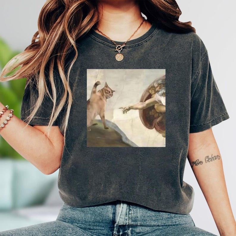 A woman wearing a gray t-shirt with a graphic design depicting a cat and a winged figure, possibly referencing the biblical scene of the creation of Adam.