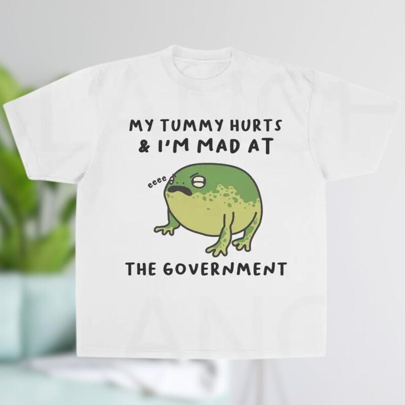 K&ouml;nnte beinhalten: Wei&szlig;es T-Shirt mit dem Text "MY TUMMY HURTS & I'M MAD AT THE GOVERNMENT". Das Shirt zeigt einen Cartoon-Frosch mit grimmigem Gesichtsausdruck. Der Frosch ist gr&uuml;n und gelb.