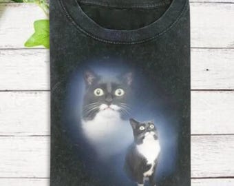 May include: Black t-shirt featuring a graphic of two cats. One cat is a close-up of the face, and the other is a full body image. Both cats have black and white markings and large, expressive eyes. The shirt has a crew neck.