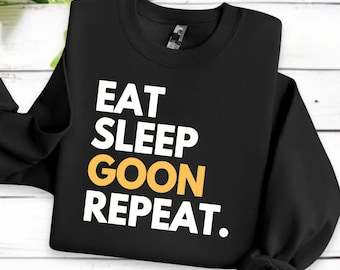 May include: Black crew neck sweatshirt with the text "EAT SLEEP GOON REPEAT" in white and gold lettering. The sweatshirt is made of a soft material and is displayed on a white wooden surface.