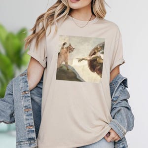 A woman wearing a beige t-shirt with a graphic of a cat and a human figure, along with a denim jacket.