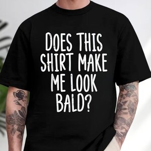May include: A black short-sleeved t-shirt featuring white text printed on the front that reads, "DOES THIS SHIRT MAKE ME LOOK BALD?" The lettering is in a casual, sans-serif font style, centered on the chest of the garment.