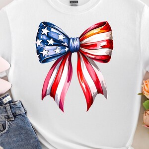 May include: White t-shirt featuring a watercolor bow design in red, white, and blue, with stars on the blue section. The shirt is paired with blue jeans and pink shoes, with a bouquet of flowers.