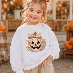 May include: A white long-sleeve sweatshirt featuring a central graphic of a pumpkin with a floral and plaid patchwork pattern, topped with a large bow. The pumpkin has a classic jack-o'-lantern face with triangular eyes and a jagged mouth.