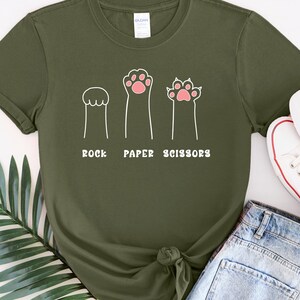 Puede incluir: Camiseta verde militar con un dise&ntilde;o l&uacute;dico de "Piedra, Papel o Tijeras" con ilustraciones de patas de dibujos animados. La camiseta se combina con pantalones cortos de mezclilla y zapatillas blancas con detalles rojos.