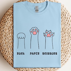 Puede incluir: Camiseta azul claro con un dise&ntilde;o l&uacute;dico de "Piedra, Papel, Tijeras" con ilustraciones de patas de gato. El texto y las almohadillas de las patas son negros y rosas. La camiseta se muestra sobre una superficie circular tejida.