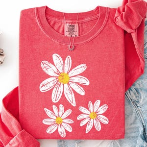 May include: A red long-sleeve shirt with a daisy graphic. The shirt is paired with denim shorts and accessories. The word "WATERMELON" is printed at the top of the image. The shirt is from Comfort Colors.