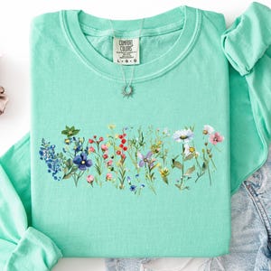 May include: A mint green long-sleeved shirt with a floral design across the chest. The design features a variety of colorful wildflowers. The shirt is folded with a pair of denim shorts and a scrunchie. The text "ISLAND REEF" is at the top.
