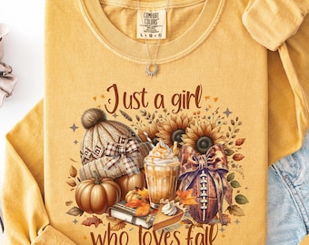 Può includere: Una maglia a maniche lunghe color giallo senape con una stampa grafica che riporta il testo Just a girl who loves fall. Il design include un berretto in maglia, una zucca, una bevanda al caffè, un pallone da football con fiocco e foglie.