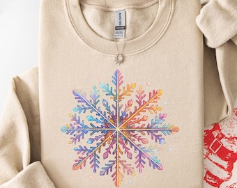 May include: A sand-colored crewneck sweatshirt featuring a large, central graphic of a multi-colored, vibrant snowflake. The snowflake design incorporates a spectrum of rainbow hues, including pink, purple, blue, orange, and yellow, with a detailed, intricate pattern. The sweatshirt has long sleeves and a ribbed collar, with a Gildan brand label visible at the inner neckline.