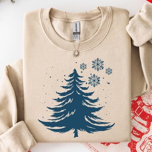 May include: A beige crewneck sweatshirt featuring a dark blue graphic of a pine tree and several snowflakes. The garment has long sleeves, a ribbed collar, and a Gildan brand label visible at the inner neckline. The design is centered on the chest.