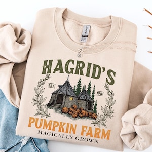 May include: A beige crewneck sweatshirt featuring a graphic print of a stone hut surrounded by pine trees and pumpkins. The text reads HAGRID'S PUMPKIN FARM, EST. 1981, and MAGICALLY GROWN. The design includes olive green and orange lettering.