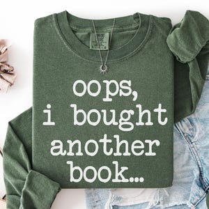 May include: An olive green long-sleeve crewneck sweatshirt featuring the white text "oops, i bought another book..." printed on the front. The garment includes a tag at the neckline and is displayed alongside a scrunchie and denim shorts.
