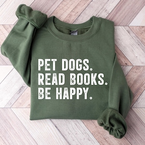 May include: A military green sweatshirt with the text "PET DOGS. READ BOOKS. BE HAPPY." in white block letters. The sweatshirt is displayed on a light wood-grain surface. The sleeve cuffs are gathered.