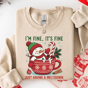 Pu&ograve; includere: Una felpa beige a girocollo con la grafica di un pupazzo di neve seduto in una tazza rossa con motivi a fiocchi di neve bianchi. Il testo recita I'M FINE, IT'S FINE e JUST HAVING A MELTDOWN. Il pupazzo di neve indossa un cappello.