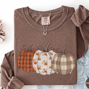 May include: A brown long-sleeve t-shirt featuring a graphic of three pumpkins with different patterns: one orange plaid, one white with orange floral motifs, and one beige checkered. The shirt has a crew neckline and a Comfort Colors brand label.