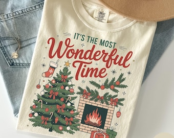 May include: An ivory t-shirt featuring the text IT'S THE MOST Wonderful Time in red and green. The graphic includes a decorated Christmas tree, a fireplace with a fire, a stocking, and wrapped gifts, all rendered in a festive holiday illustration.