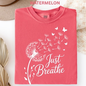 May include: Watermelon-colored Comfort Colors t-shirt with a white dandelion design and the words "Just Breathe." The design includes butterflies and a floral element. The shirt is a crew neck.