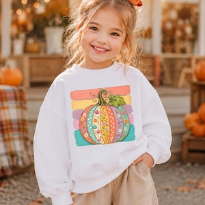 May include: A white long-sleeve sweatshirt featuring a central graphic of a pumpkin decorated with colorful floral patterns and polka dots, set against a background of horizontal pastel-colored brushstrokes in pink, yellow, and teal.