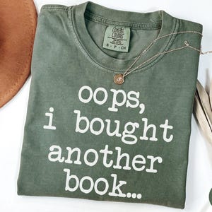 May include: A moss green short-sleeved t-shirt with a crew neckline and the white text "oops, i bought another book..." printed on the front. The shirt features a Comfort Colors brand label on the inside collar and is size small.