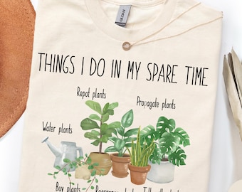 Può includere: Una t-shirt color naturale con il testo "THINGS I DO IN MY SPARE TIME" sopra un'illustrazione ad acquerello di varie piante in vaso e un annaffiatoio, con etichette come "Water plants," "Repot plants," "Propagate plants," "Buy plants," "Rearrange plants," e "Talk with plants."