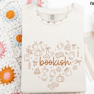 A white t-shirt with a heart-shaped design featuring the word "bookish" and various fall-themed illustrations, such as pumpkins, leaves, and mugs, is displayed on a white surface.