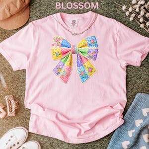 May include: Pale pink t-shirt with a vibrant patchwork bow design. The bow incorporates floral and geometric patterns in pink, blue, yellow, and green. The word "BLOSSOM" is printed at the top. The shirt is made of soft material.