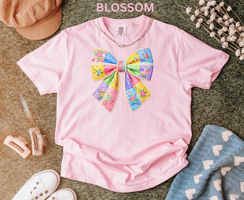 May include: Pale pink t-shirt with a vibrant patchwork bow design. The bow incorporates floral and geometric patterns in pink, blue, yellow, and green. The word "BLOSSOM" is printed at the top. The shirt is made of soft material.