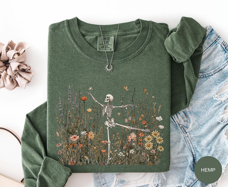Puede incluir: Una sudadera de manga larga color verde bosque con un estampado gr&aacute;fico de un esqueleto bailando entre flores silvestres coloridas y mariposas. La prenda tiene cuello redondo y una etiqueta de la marca Comfort Colors en el cuello interior. Un collar con colgante de sol plateado descansa sobre el pecho.