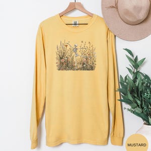Puede incluir: Una camiseta de manga larga color amarillo mostaza con un estampado gr&aacute;fico de un esqueleto en una postura de yoga rodeado de flores silvestres y vegetaci&oacute;n. La prenda tiene cuello redondo y se muestra en una percha de madera.