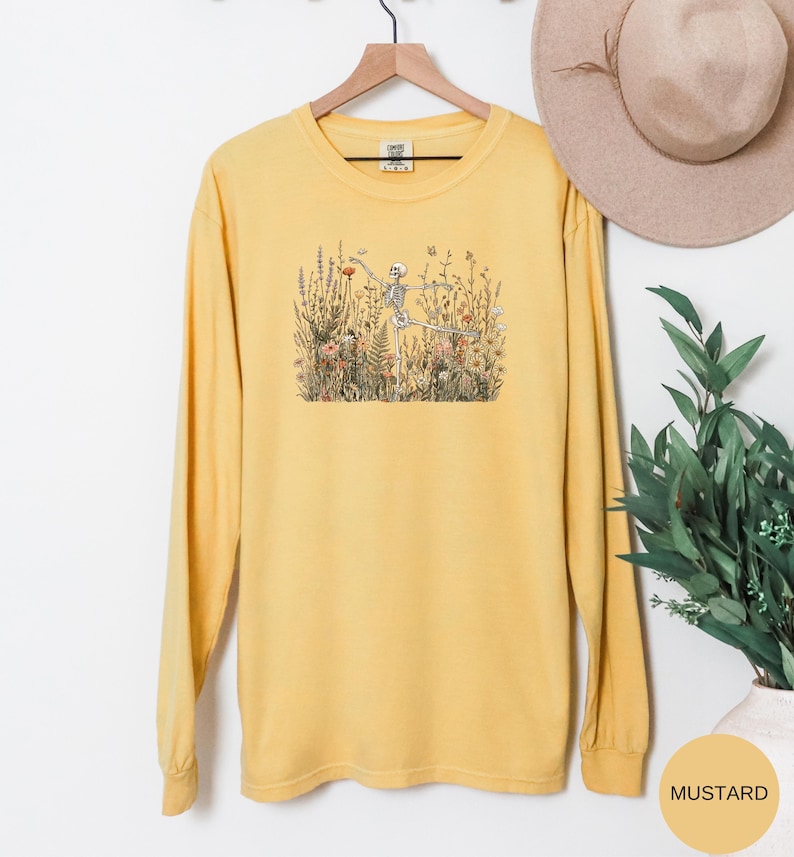Puede incluir: Una camiseta de manga larga color amarillo mostaza con un estampado gr&aacute;fico de un esqueleto en una postura de yoga rodeado de flores silvestres y vegetaci&oacute;n. La prenda tiene cuello redondo y se muestra en una percha de madera.