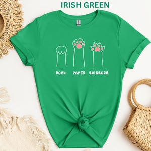 Puede incluir: Camiseta verde irland&eacute;s con un dise&ntilde;o l&uacute;dico que presenta ilustraciones de patas de gato que representan piedra, papel y tijeras. El texto "ROCK PAPER SCISSORS" est&aacute; impreso debajo. La camiseta est&aacute; anudada a la cintura.