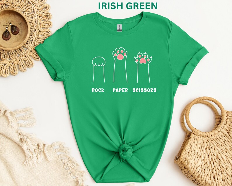 Puede incluir: Camiseta verde irland&eacute;s con un dise&ntilde;o l&uacute;dico que presenta ilustraciones de patas de gato que representan piedra, papel y tijeras. El texto "ROCK PAPER SCISSORS" est&aacute; impreso debajo. La camiseta est&aacute; anudada a la cintura.