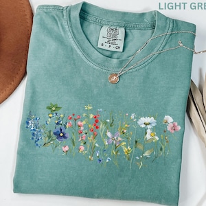 May include: Light green t-shirt with a floral design across the front. The shirt has a crew neck and short sleeves. A silver necklace with a round pendant rests on the shirt. The shirt is folded and has a tag that says "Comfort Colors".