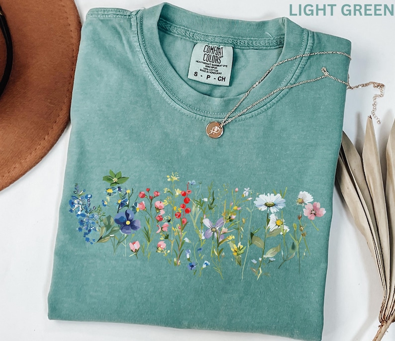 May include: Light green t-shirt with a floral design across the front. The shirt has a crew neck and short sleeves. A silver necklace with a round pendant rests on the shirt. The shirt is folded and has a tag that says "Comfort Colors".