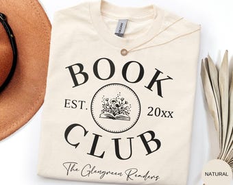 May include: A cream-colored short-sleeve t-shirt featuring black text that reads BOOK CLUB, EST. 20xx, and The Glengreen Readers. A central circular graphic displays an open book with flowers emerging from the pages.