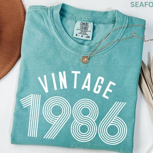 May include: Seafoam green t-shirt with the words "VINTAGE 1986" in white block letters. The shirt has a crew neck and short sleeves. A gold necklace with a pendant is draped across the shirt.