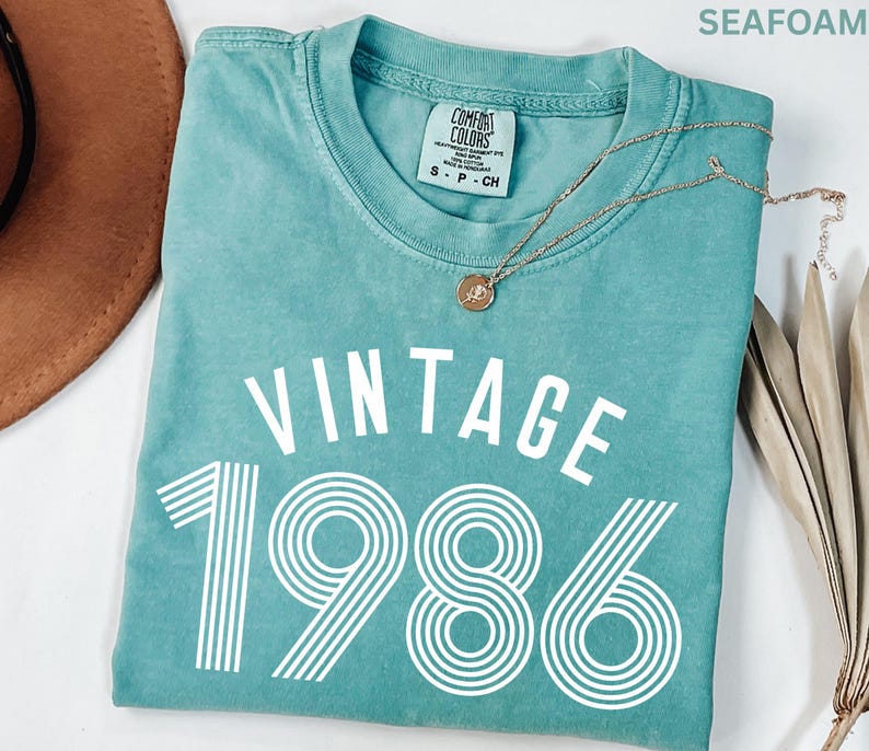 May include: Seafoam green t-shirt with the words "VINTAGE 1986" in white block letters. The shirt has a crew neck and short sleeves. A gold necklace with a pendant is draped across the shirt.