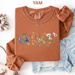 May include: A burnt orange long-sleeve shirt with a crew neck, featuring a floral design across the chest. The design includes blue, red, and white flowers. The word "YAM" is in the top left corner.