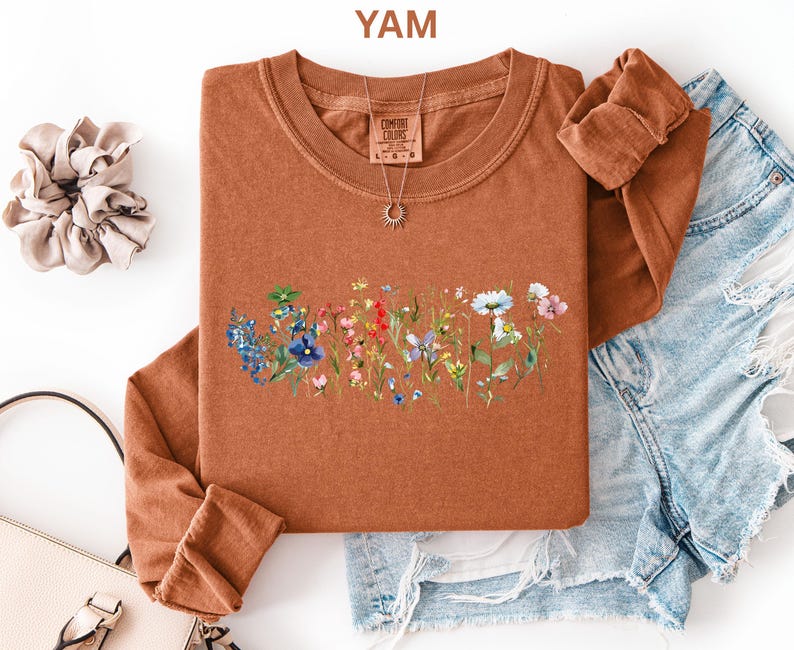 May include: A burnt orange long-sleeve shirt with a crew neck, featuring a floral design across the chest. The design includes blue, red, and white flowers. The word "YAM" is in the top left corner.