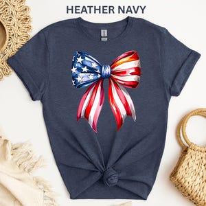 May include: Heather navy t-shirt featuring a watercolor bow design in red, white, and blue, with stars on the blue section. The shirt is knotted at the waist. The text "HEATHER NAVY" is at the top.
