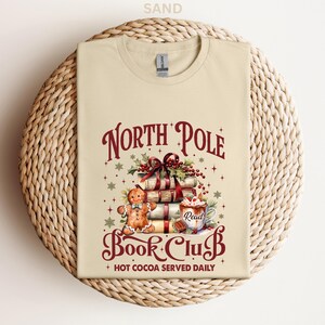 A beige t-shirt with a Christmas-themed design, including a gingerbread man, books, and a hot chocolate mug, along with the text "North Pole Book Club" and "Hot Cocoa Served Daily".