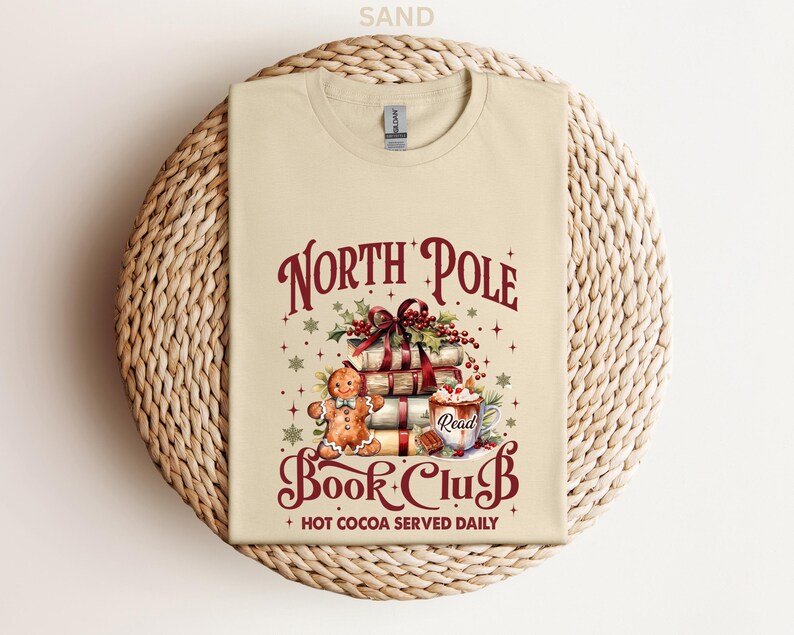 A beige t-shirt with a Christmas-themed design, including a gingerbread man, books, and a hot chocolate mug, along with the text "North Pole Book Club" and "Hot Cocoa Served Daily".
