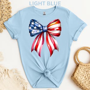 May include: Light blue t-shirt featuring a watercolor bow design in red, white, and blue, with stars and stripes. The shirt is knotted at the waist. The text "LIGHT BLUE" is at the top.