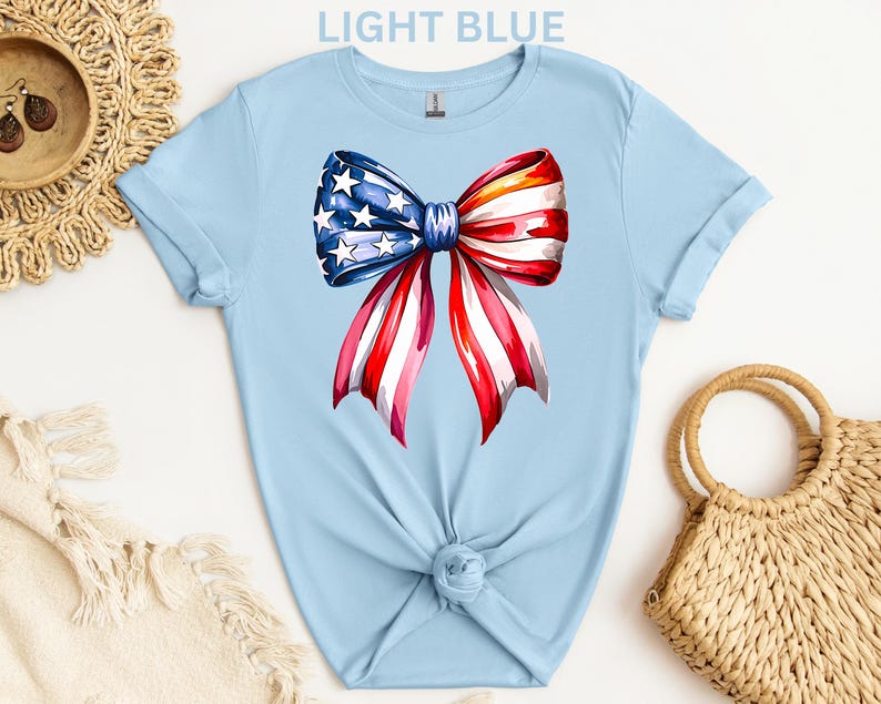 May include: Light blue t-shirt featuring a watercolor bow design in red, white, and blue, with stars and stripes. The shirt is knotted at the waist. The text "LIGHT BLUE" is at the top.