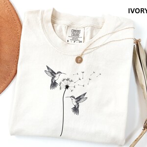 a white t-shirt with a black and white illustration of two hummingbirds flying next to a dandelion, along with a necklace and a hat.