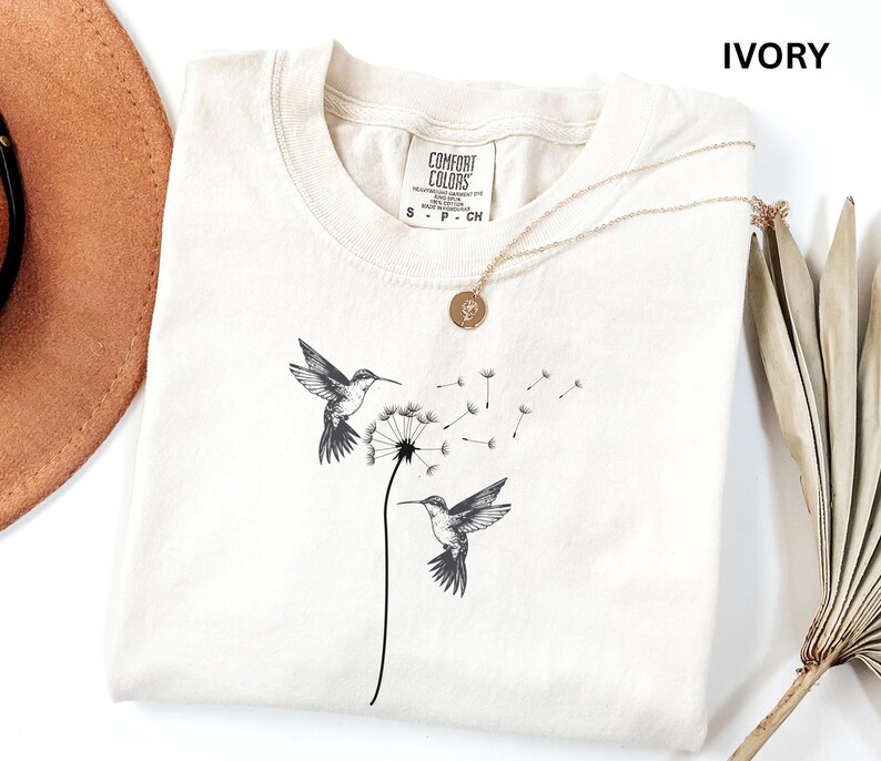 a white t-shirt with a black and white illustration of two hummingbirds flying next to a dandelion, along with a necklace and a hat.
